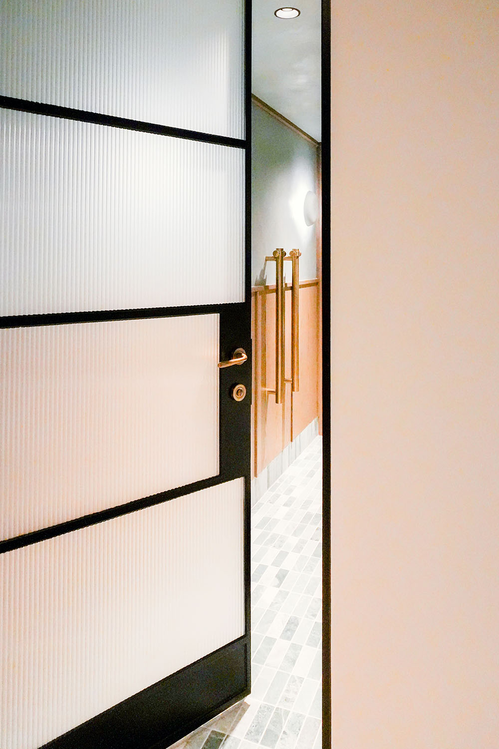 Black steel-framed glass door partially open, revealing a stylish interior with textured walls and elegant brass handles.