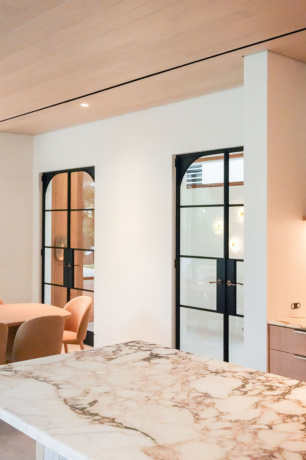 Elegant steel frame glass hinge doors in a modern interior, featuring a marble island and soft-toned furnishings, enhancing the space's design.
