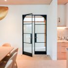 Double glass hinge doors with a black steel frame open into a minimalist space, featuring light wood and marble surfaces.