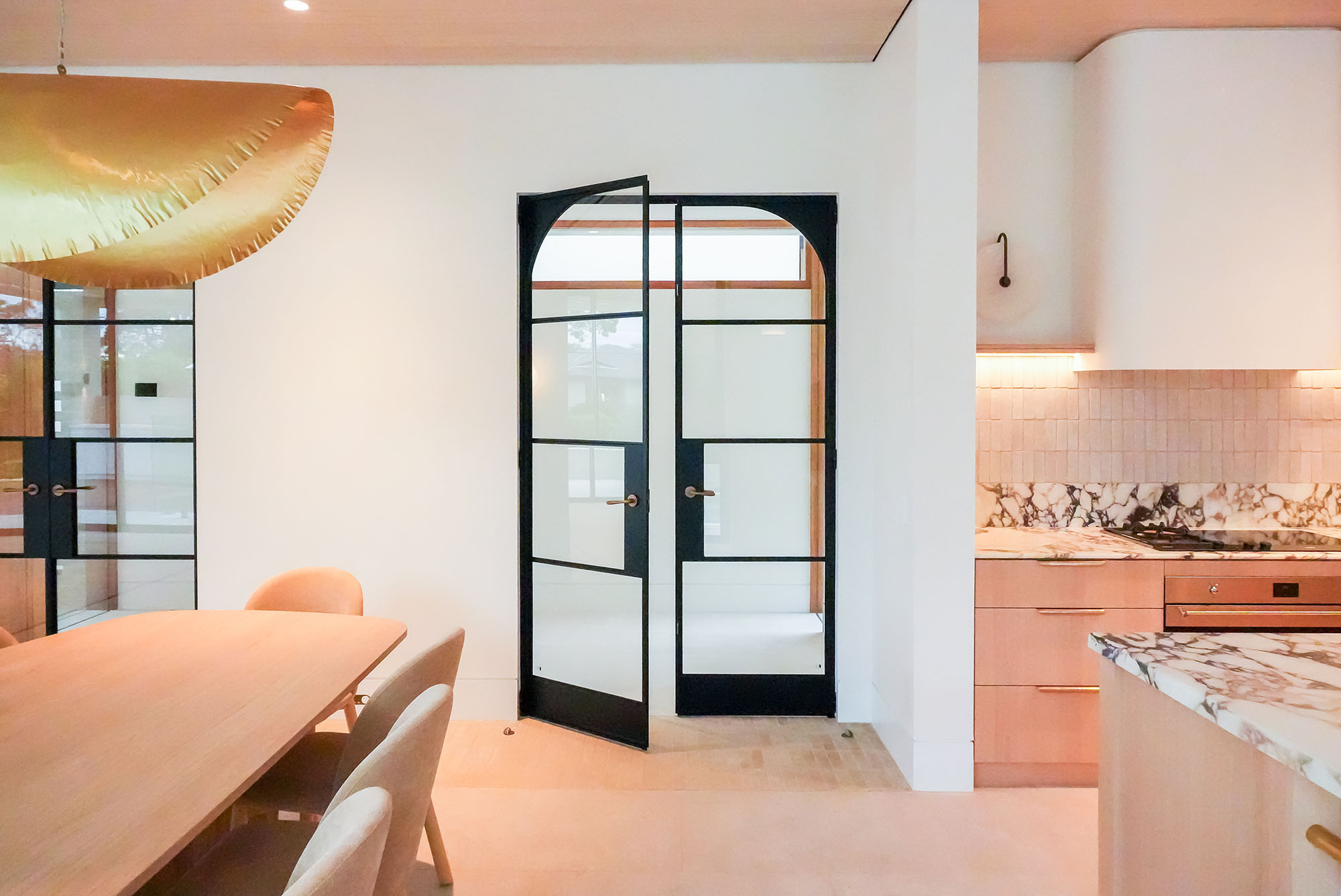 Double glass hinge doors with a black steel frame open into a minimalist space, featuring light wood and marble surfaces.