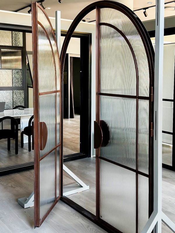 Bespoke steel doors with arched tops and ribbed glass panels in a modern showroom, featuring a light wood floor and reflective surfaces.