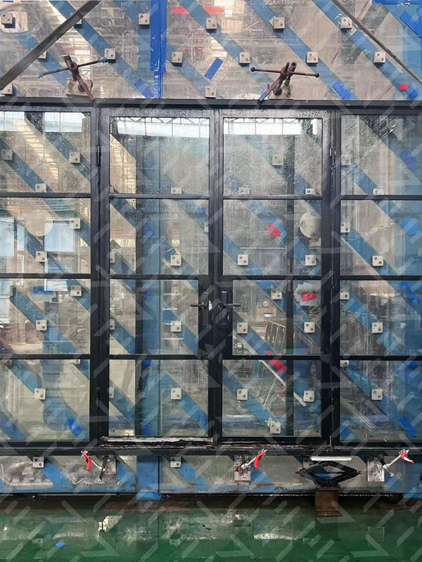Black steel-framed windows with multiple glass panes, set against a backdrop of industrial testing equipment and blue supports.