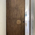 Textured iron pivot door with vertical grooves, featuring a circular handle, set in a light-toned wall. Natural light filters through.