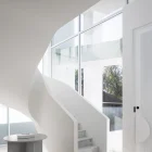 Curved white staircase leads to large windows in a minimalist interior, featuring soft light and a round marble table.