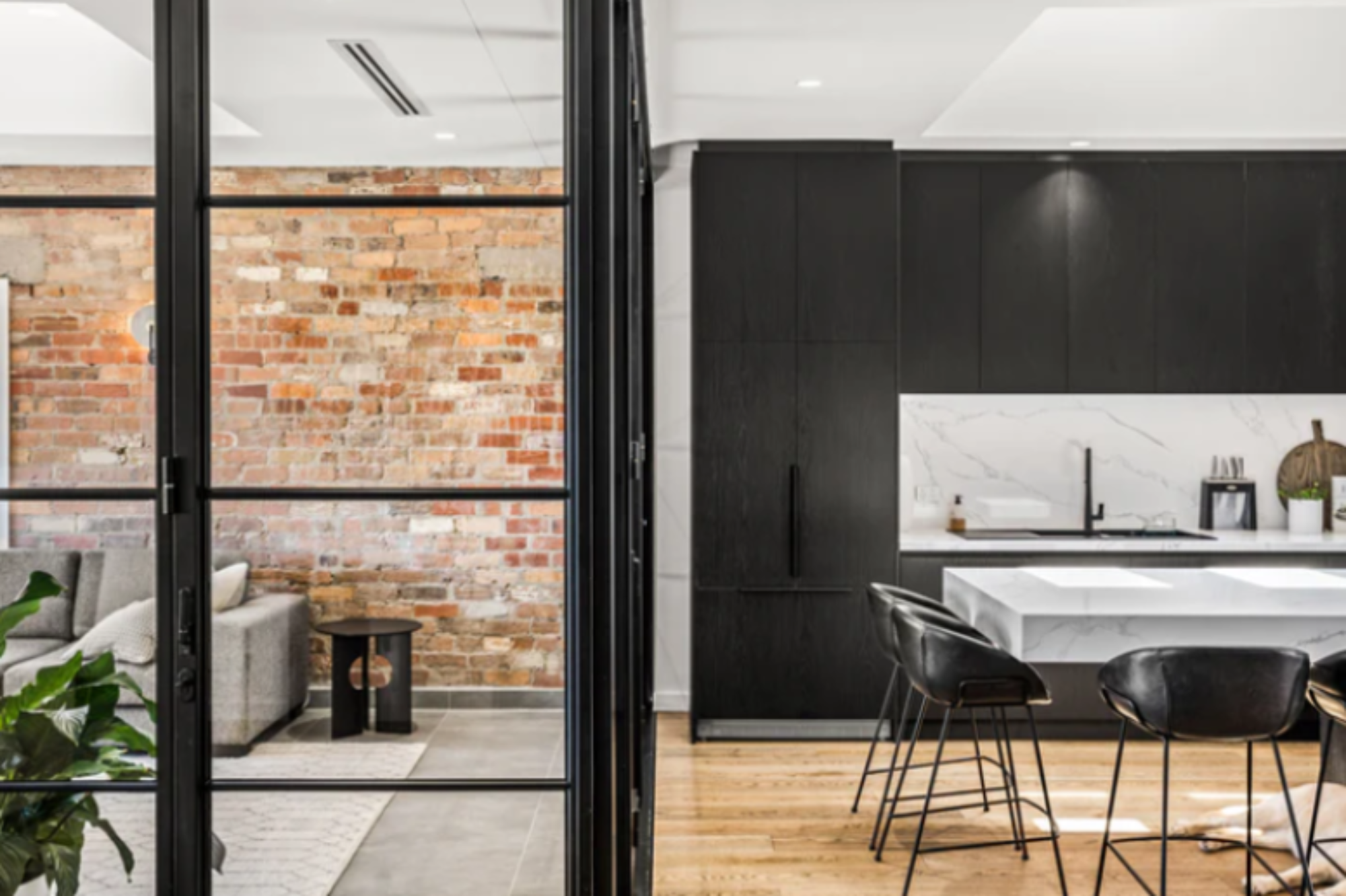 Black steel-framed fixed windows divide a modern living area with exposed brick and a sleek kitchen featuring marble countertops and bar stools.