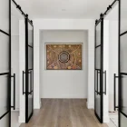 Two black steel frame sliding doors open into a hallway, showcasing a circular artwork against a neutral wall, with light wooden flooring.