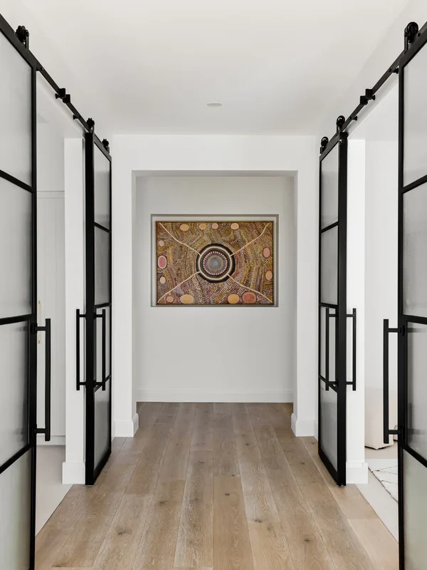 Two black steel frame sliding doors open into a hallway, showcasing a circular artwork against a neutral wall, with light wooden flooring.
