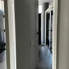 Modern iron pivot entry door with vertical ribbing, framed in white, opening into a sleek, light-filled interior space with wooden flooring.