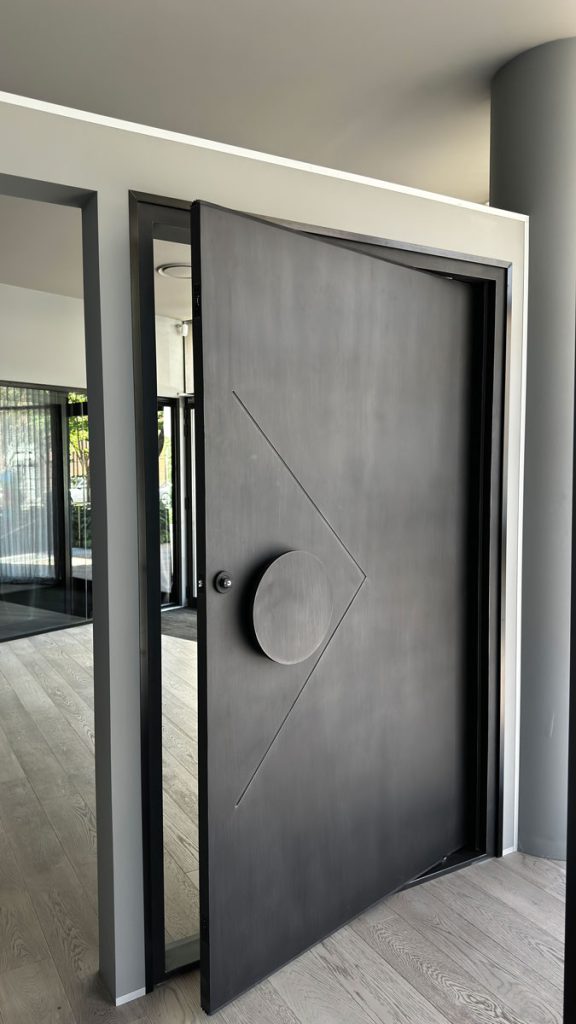 Bespoke Architectural Steel Door Design by Zen Doors & Windows A sleek black architectural steel door with a circular handle, partially open, framed by a minimalist grey wall and light wooden flooring.