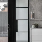 A sleek black steel-framed glass door with ribbed panels, contrasting against a light grey stone wall and a modern kitchen area.