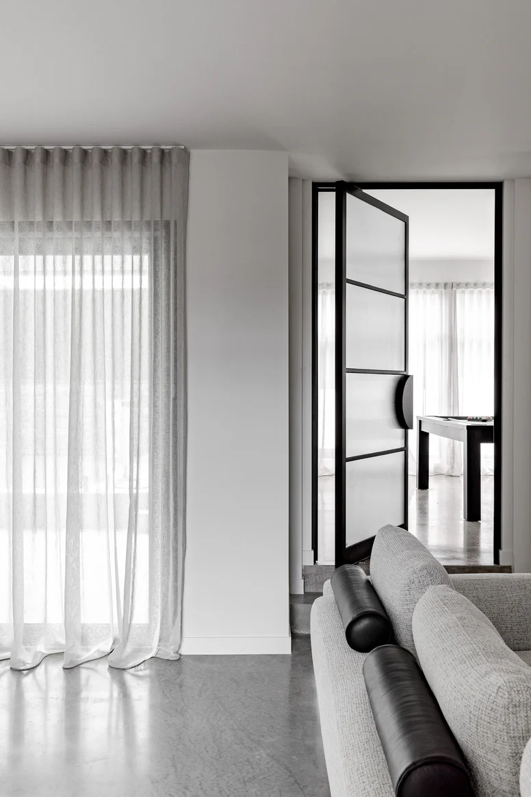 Glass pivot door with a sleek black frame, open to a light-filled room with sheer curtains and a modern sofa in grey tones.