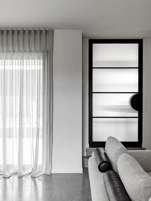 Modern interior featuring a textured black glass pivot door beside sheer grey curtains and a light grey sofa, creating a minimalist aesthetic.