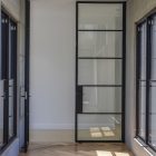 Steel-framed glass hinge door in a modern interior, flanked by large windows, with herringbone wooden flooring and soft lighting.