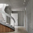 Curved wooden staircase leads to a light-filled corridor with concrete walls and a white panel door, highlighting modern architectural simplicity.