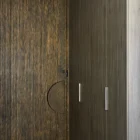 Textured iron batten hinge door with a circular handle, set against muted wood paneling and a sleek floor, creating a modern aesthetic.