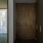 Textured dark bronze entry door with curved cutout, set in a minimalist hallway with wood and glass elements, soft light filtering through.