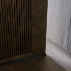 Close-up of a dark bronze batten hinge door featuring vertical grooves, set against a pale wooden background and dark wooden flooring.