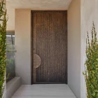 Bespoke iron entry door with a textured bronze finish, framed by tall green plants, set against a light-coloured wall and steps.