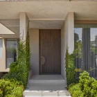 Modern entrance featuring a wooden door flanked by lush greenery, with a sleek concrete pathway leading to a minimalist facade.