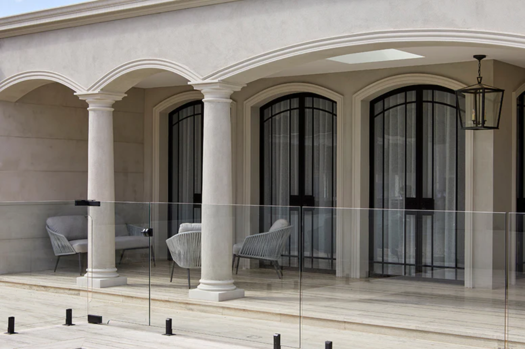 Elegant porch featuring arched columns, black-framed glass doors, and grey wicker chairs, enhanced by a glass balustrade.