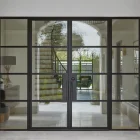 Bespoke black steel-framed glass doors with multiple panes, opening to a stylish interior featuring stairs and a garden view.