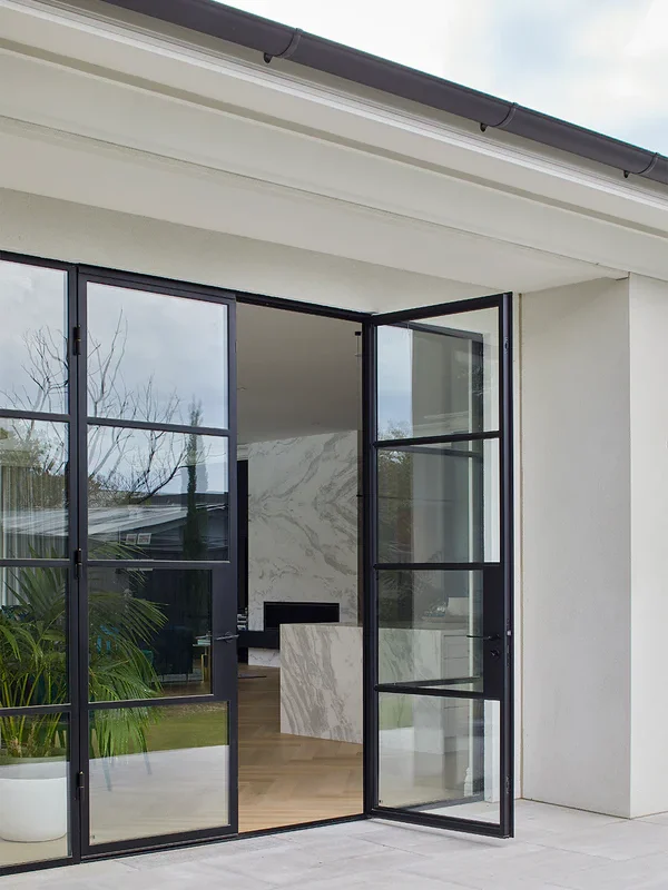 Black steel-framed glass doors open to a modern interior with marble features and greenery, set against a light-coloured façade.