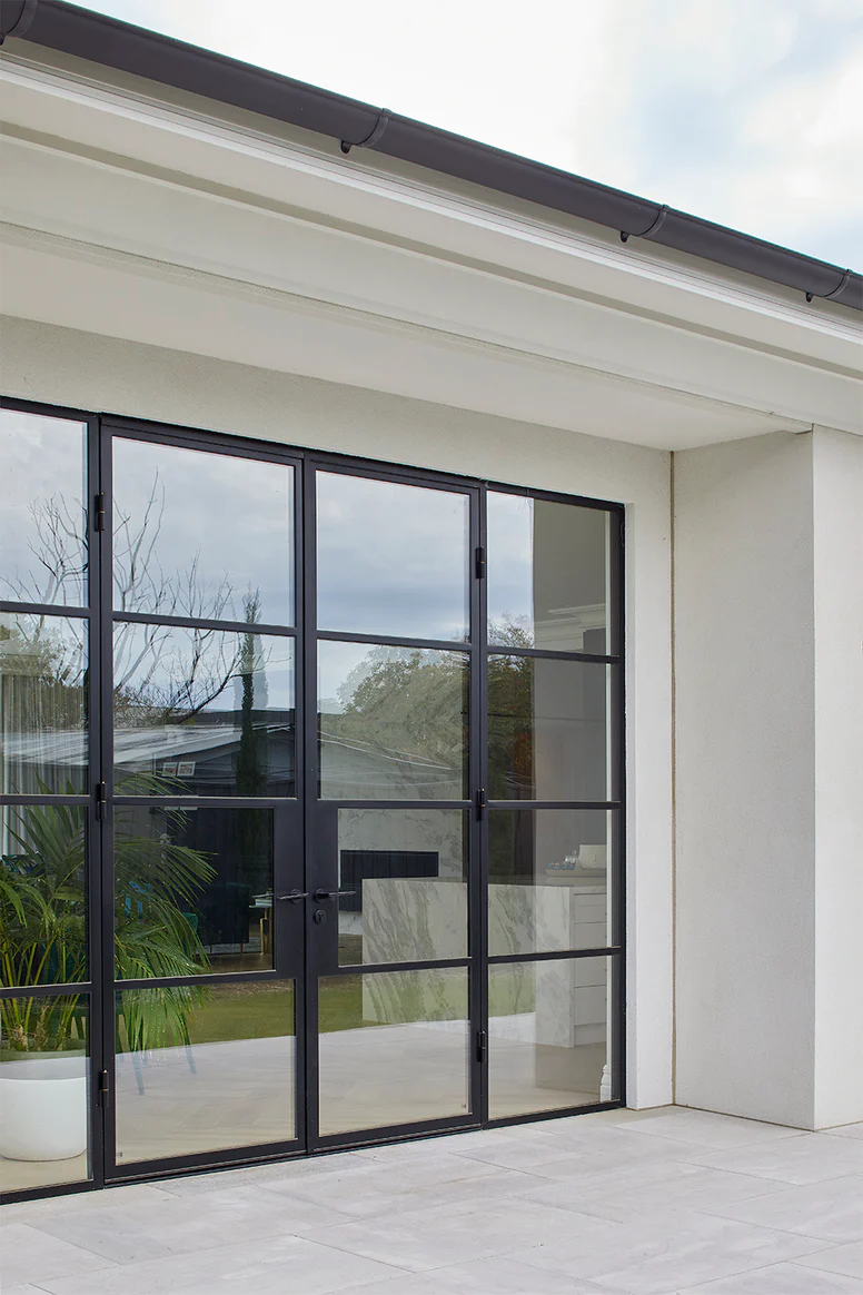 Bespoke black steel-framed glass doors with multiple panes, set into a modern white building, featuring a patio and greenery beyond.