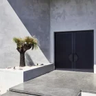 Elegant black iron batten hinge doors framed by a modern, minimalist concrete façade, featuring a landscaped entry with a grassy tree.