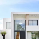 Bespoke white modern house featuring a large black iron entry door, large windows, and landscaped greenery under a blue sky.