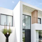 Modern white house façade featuring large black entry door, vertical wooden slats, and landscaped garden with plants and unique tree.