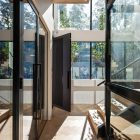 Dark iron pivot door opens into a light-filled space with large glass walls, showcasing greenery and wooden stairs in a contemporary design.