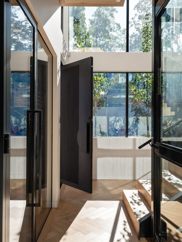 Dark iron pivot door opens into a light-filled space with large glass walls, showcasing greenery and wooden stairs in a contemporary design.