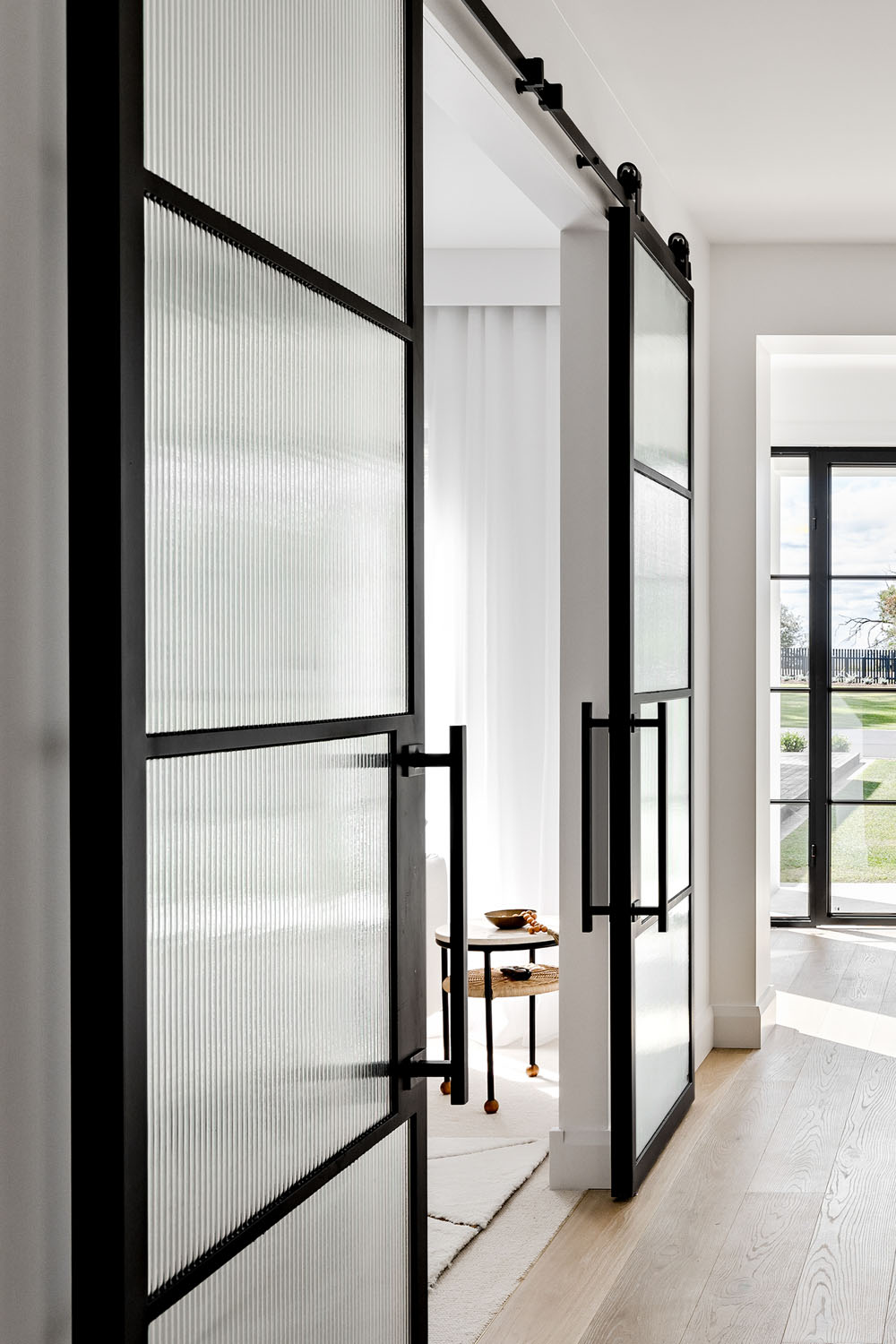 Internal view of sleek galvanised steel frame glass barn sliding doors, featuring textured glass panels and modern black handles, framed by light wood flooring.