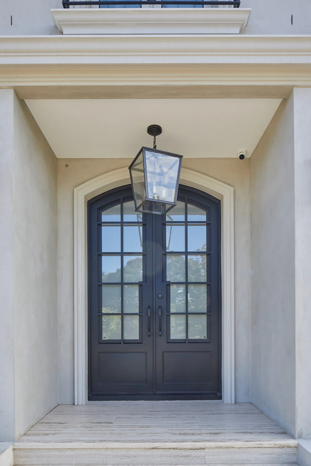 Elegant double-entry door with black wrought iron hinges and glass panels, flanked by a stylish lantern, set against a creamy stone façade.