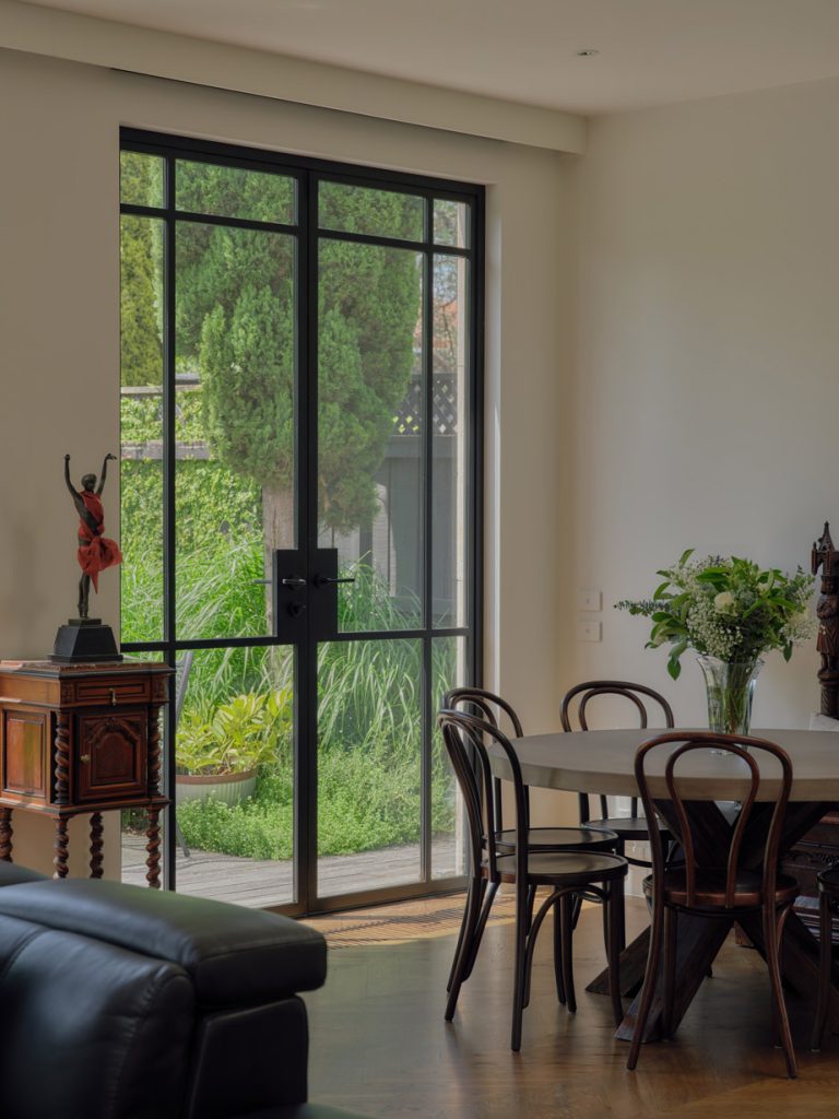 Black steel-framed glass doors open to a lush green garden, with a wooden dining table and vintage decor in a modern living space.