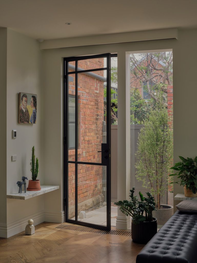 Black steel-framed door opens to a lush courtyard; interiors feature earthy tones, plants, and elegant wood flooring.