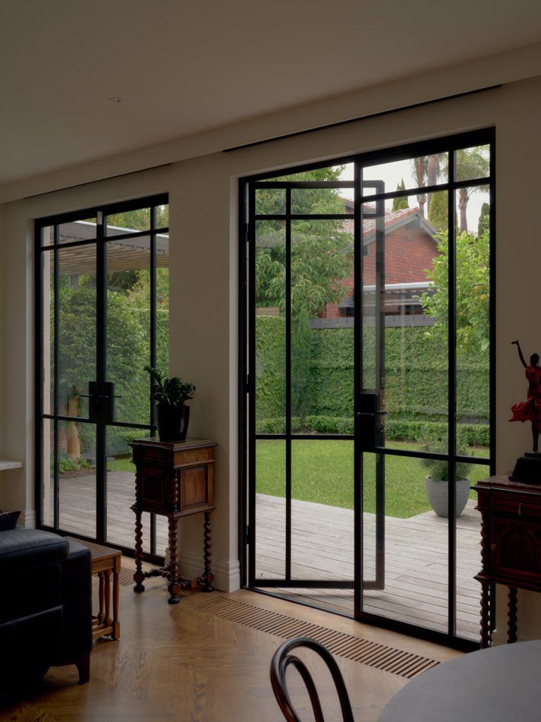 Bespoke black steel-framed doors open to a lush green garden, featuring wooden flooring, potted plants, and natural light streaming in.