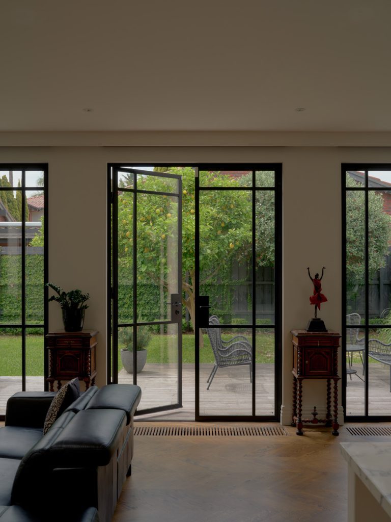 Elegant black steel-framed doors open to a lush garden, featuring greenery and outdoor seating, set in a modern interior with stylish furnishings.