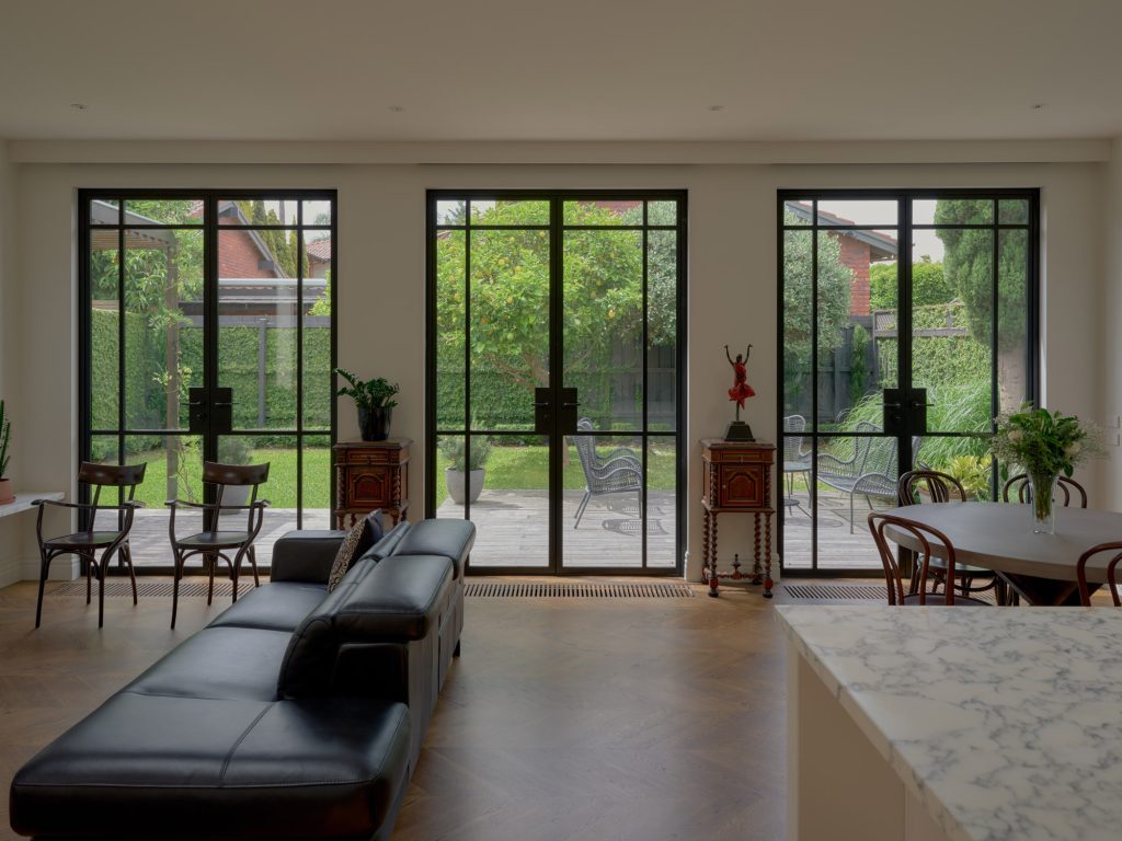 Modern living space featuring a black leather sofa, wood dining area, and expansive steel-framed windows overlooking a landscaped garden.