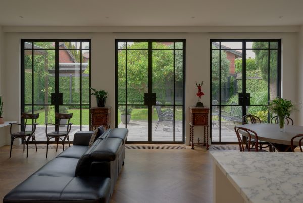 Modern living space featuring a black leather sofa, wood dining area, and expansive steel-framed windows overlooking a landscaped garden.
