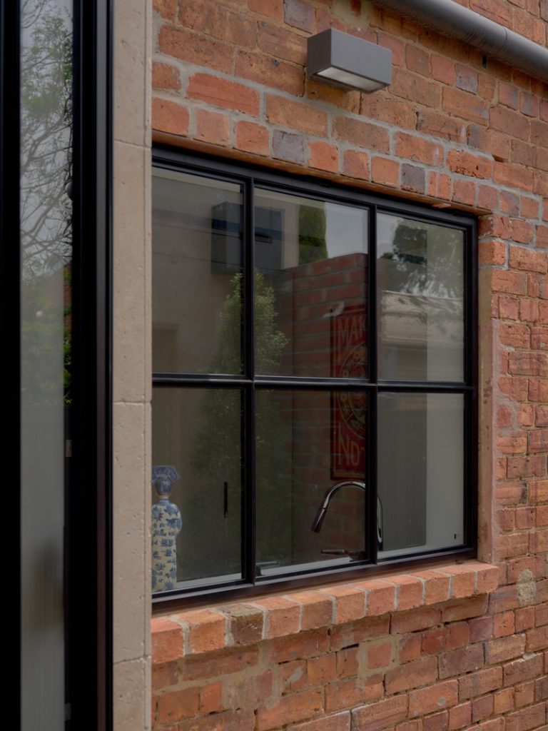 Black steel-framed window set within a textured brick wall, showcasing a modern design and a glimpse of greenery outside.