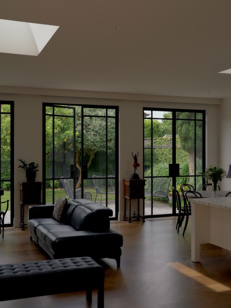 Bright living space featuring large steel-framed windows, a modern black leather sofa, and views of a lush garden outside.