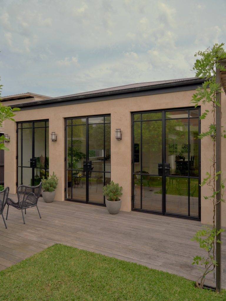 Bespoke steel-framed glass doors on a limestone house, surrounded by planters, a wooden deck, and lush greenery under a cloudy sky.