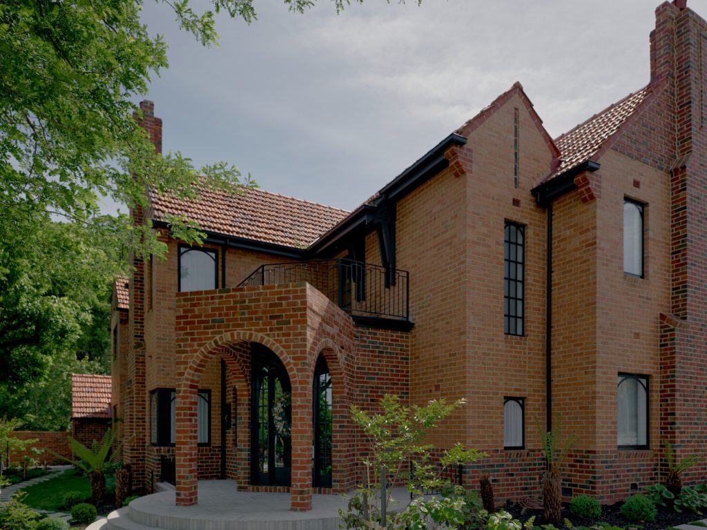 Red-brick house with a tiled roof, featuring arched entrance, large windows, and surrounding greenery, showcasing elegant architectural design.