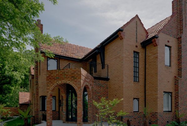 Red-brick house with a tiled roof, featuring arched entrance, large windows, and surrounding greenery, showcasing elegant architectural design.