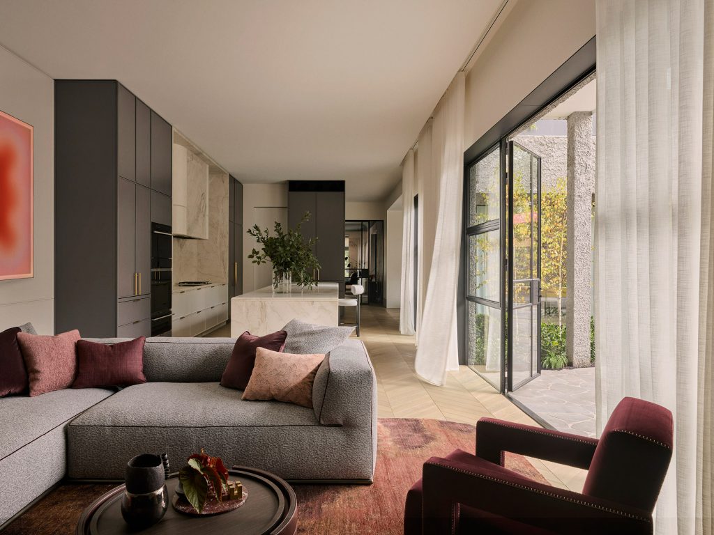 Modern lounge featuring a grey modular sofa, burgundy accents, and large glass doors opening to a landscaped outdoor area.