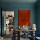 A vibrant blue office features a striking orange artwork above a stylish desk, complemented by modern furniture and elegant decor elements.