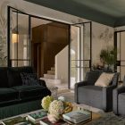 Elegant living space featuring dark velvet sofa, grey armchairs, and a glass coffee table stacked with books. Botanical wallpaper and steel-framed doors enhance the design.