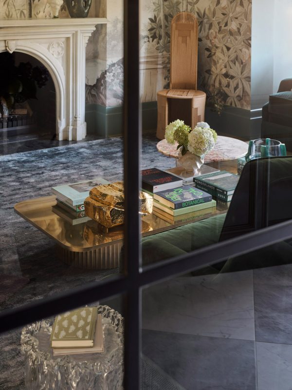 A stylish interior featuring a marble fireplace, elegant furniture, and a coffee table stacked with books, surrounded by floral wallpaper.