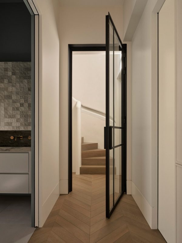 A black steel-framed glass door opens into a light hallway with a herringbone wooden floor, leading to a staircase beyond.